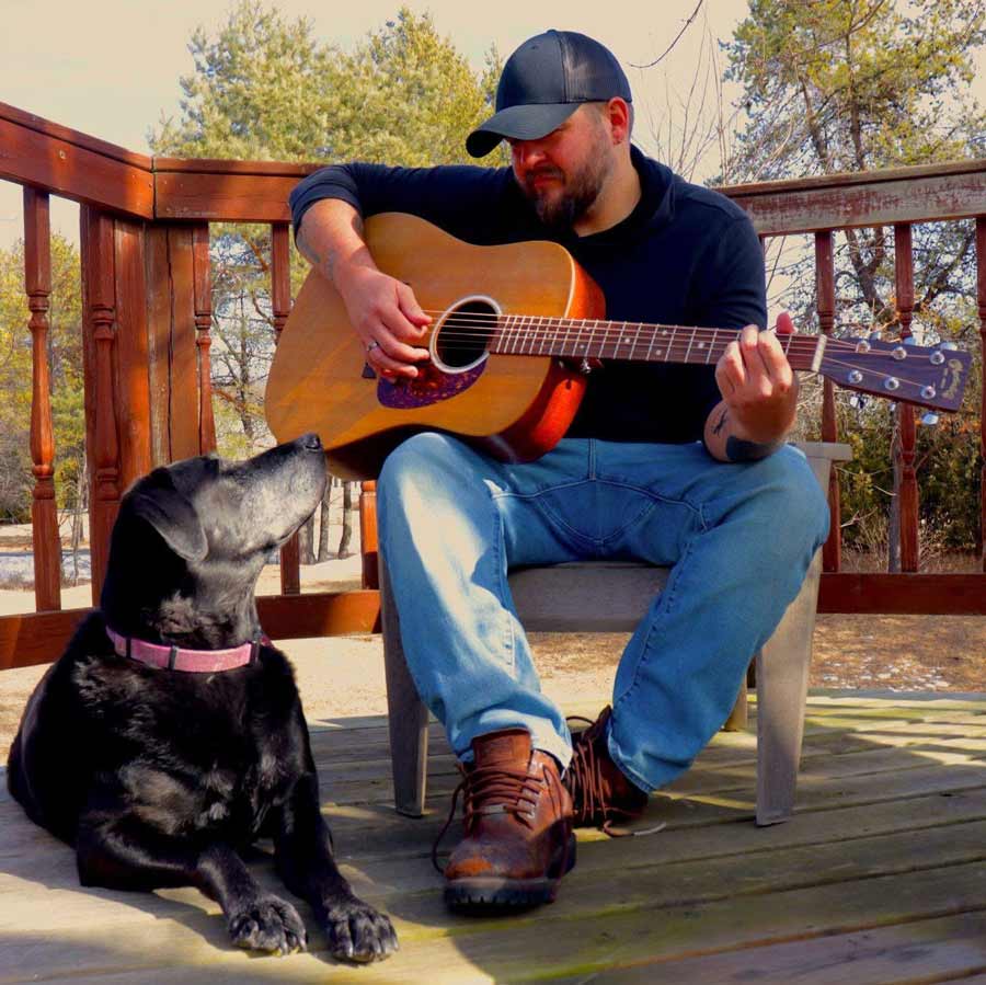Ryders Roadhouse in Wisconsin Rapids Wisconsin Man playing guitar on a porch while a black dog lies nearby, looking up at him.
